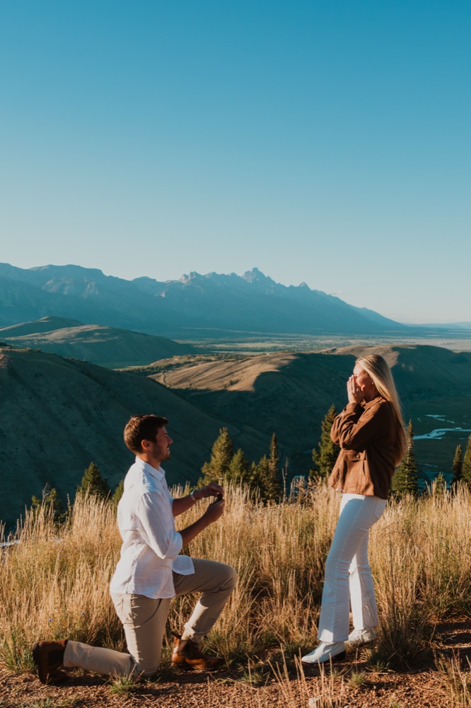 Alexander kneeling to propose to Julia with the Teton range behind them